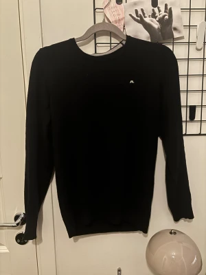 Svart stickad tröja från J.Lindeberg - Svart stickad tröja från J.Lindeberg med diskret logga på bröstet. Klassisk rund hals och långärmad modell, perfekt för dig som gillar stilrena och enkla plagg. Mjuk och skön kvalitet som passar till många olika looks. 