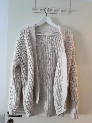 Beige stickad kofta med grov struktur - Säljer en beige stickad kofta med grov struktur och öppet framtill. Koftan har långärmade ärmar och breda ribbade muddar vid ärmslut och nederkant. Perfekt för att slänga över en t-shirt när det är lite kyligt ute.