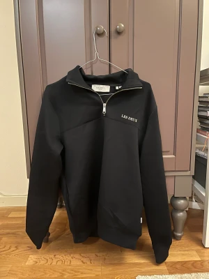 Mörkblå  half zip tröja från Les Deux - Mörkblå långärmad tröja från Les Deux med half zip-dragkedja och broderad logga på bröstet. Tröjan har en hög krage och är tillverkad i mjukt material som ger en clean och stilren look. Perfekt för dig som gillar minimalistisk design.