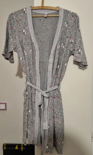 Grå blommig morgonrock med bälte - Säljer en grå kortklänning med blommigt mönster i rosa, vitt och svart. Klänningen har korta ärmar, omlott-design och ett randigt bälte i midjan. Materialet är mjuk bomull och kanten har diskreta ränder. Perfekt för dig som gillar chill och söt stil. Storlek S/M