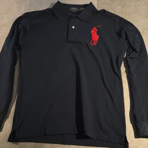 Marinblå långärmad pikétröja Polo Ralph Lauren - Snygg marinblå långärmad pikétröja från Polo Ralph Lauren med röd broderad logga på bröstet och en stor röd siffra 3 på ärmen. Klassisk krage och knappar framtill. Tröjan är i bomull och har en slim passform som sitter riktigt schysst.