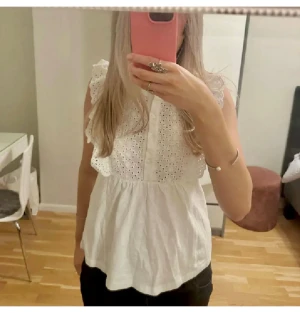 Vit broderad blus från JDY i stl XS - Supersöt vit blus från JDY i storlek XS. Blusen är ärmlös med volangkant vid axlarna, knappar framtill och broderade hålmönster över bröstet. Tillverkad i bomull, perfekt för en fräsch och somrig look. Se lite sminkfläckar på bild 5.
