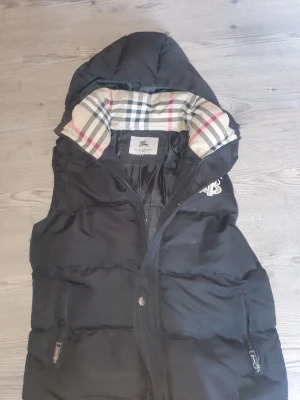 Svart dunväst från Burberry med huva - Svart dunväst från Burberry med ikonisk rutig insida i beige, svart och rött. Västen har huva, dragkedja och två fickor med dragkedja. Broderad Burberry-logga på bröstet. Perfekt för lager-på-lager och snygg streetstil.