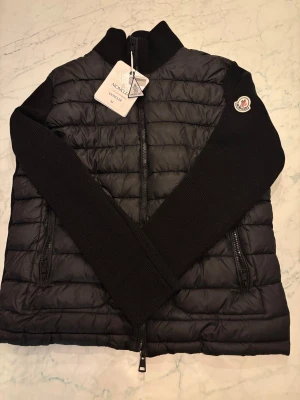 Svart quiltad Moncler jacka M - Svart quiltad jacka från Moncler i storlek M. Jackan har stickade ärmar och krage, två dragkedjefickor och Moncler-logga på ärmen. Snygg, stilren design med ribbade detaljer och hel dragkedja framtill. Perfekt för dig som gillar exklusiva märken och modern look.