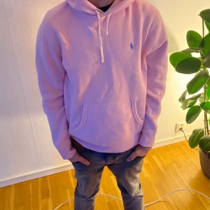 Rosa hoodie från Ralph Lauren - Säljer en ljusrosa hoodie från Ralph Lauren med klassisk huva och snörning. Tröjan har en känguruficka framtill och den ikoniska blå loggan broderad på bröstet. Perfekt för dig som gillar en avslappnad och stilren look. Liten liten defekt med ett hål man knappt ser och även fläck som är borttvättad som knappast syns 