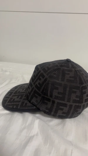 Svart Fendi keps med FF-mönster - Snygg svart keps från Fendi med det klassiska FF-logomönstret över hela kepsen. Justerbar baktill med Fendi Roma-detalj. Tillverkad i Italien och gjord av ett slitstarkt tygmaterial. Perfekt för dig som gillar lyxiga streetwear-accessoarer.