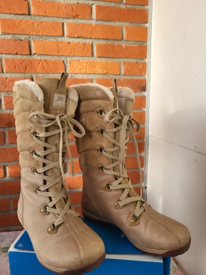 Beige Helly Hansen vinterstövlar - Snygga beige vinterstövlar från Helly Hansen med högt skaft och snörning hela vägen upp. Utsidan är i mocka och skinn, fodrade med mjuk syntetpäls för extra värme. Perfekt för kalla dagar och har en platt sula med rund tå. Vattentäta och robusta.