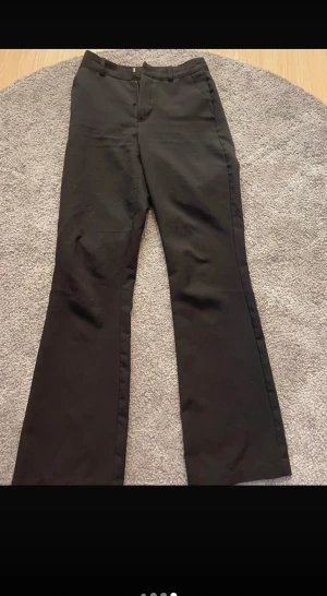 Svarta bootcut kostymbyxor - Snygga svarta kostymbyxor med bootcut-snitt och klassisk midja. Byxorna har bälteshällor, diskreta fickor bak och stängs med dragkedja och knapp framtill. Perfekta för en clean och stilren look.