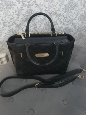 Svart handväska från Guess med axelrem - Stilren svart handväska från Guess med präglat mönster och guldfärgade detaljer. Väskan har både handtag och avtagbar axelrem, samt dragkedja upptill. Insidan är ljus med Guess-logga och har flera fack för smart organisering. Perfekt storlek för allt du behöver.