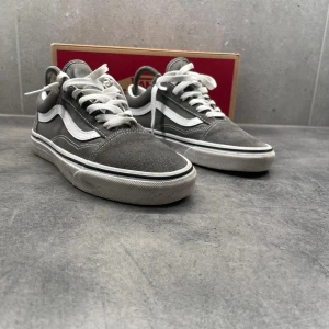 Gråa sneakers från Vans - Säljer ett par klassiska gråa Vans sneakers med vit sula och snörning. Skorna har den ikoniska vågiga sidodetaljen i vitt och en brun gummisula. Perfekta för en avslappnad stil.