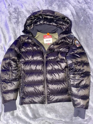 Svart pufferjacka Parajumpers Sheen - Yunior small passar 1,45-1,55 cm lång ,Snygg svart pufferjacka från Parajumpers, modell Sheen. Jackan har huva, ribbade muddar och coolt patch på ärmen. Yttermaterialet är glansigt och jackan har dragkedja framtill. Perfekt för dig som vill ha en varm och stilren look.