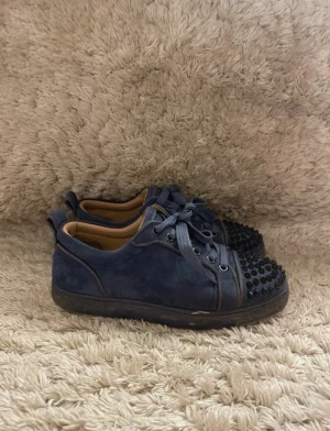 Blå Christian Louboutin - Säljer ett par blå sneakers från Christian Louboutin i mocka med coola svarta nitar på tån och klassisk röd sula. Skorna har rund tå, platt sula och snörning. Insidan är beige och de har en lyxig vibe som sticker ut.