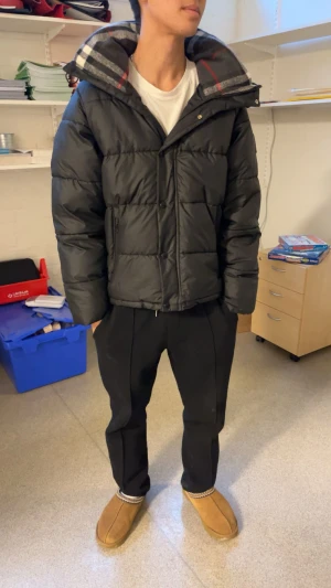 Svart Burberry pufferjacka med huva - Säljer en svart pufferjacka från Burberry med stor huva och klassiskt rutigt foder. Jackan har dragkedja och knappar framtill, samt två sidofickor med dragkedja. Märket är broderat på ärmen och jackan har en loose passform. Perfekt för kalla dagar. Köpt för några dagar sen, använd runt 5 gånger. Priset är inte hugget i sten.