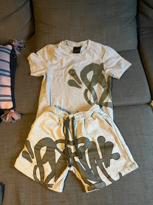 Grå syna world t-shirt och shorts  - Säljer ett par grå shorts och t-shirt med coolt grafiskt tryck i mörkgrått framtill. De har elastisk midja med snörning och en bakficka med dragkedja som har en unik zip-puller. Materialet är mjuk bomull, perfekt för chill dagar. Passformen är loose och de har en modern streetwear-vibe.