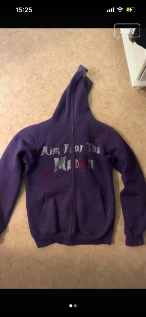 Lila hoodie Aim For The Moon  - lila zip-hoodie från Aim For The Moon i storlek L men sitter som M. Hoodien har trycket 'Aim For The Moon' i silver och röda stenar framtill. Materialet är mjukt och fleece-liknande innuti.