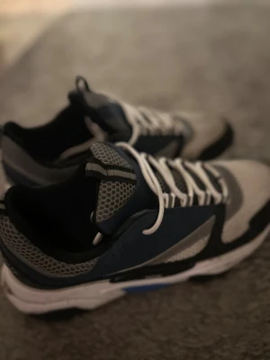 Blå och grå sneakers med meshdetaljer - Snygga sneakers i blått, grått och svart med vit sula och vita snören. Ovandelen har meshdetaljer och ett coolt hexagonmönster på tungan och sidorna. Skorna har en sportig vibe och passar perfekt för dig som gillar streetstyle.