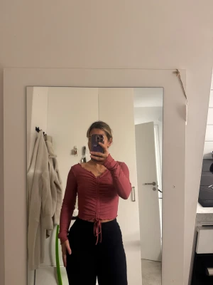 Rosa croppad topp med snörning - Säljer en rosa croppad topp med lång ärm och snygg v-ringning. Toppen har snörning framtill som ger en cool rynkad effekt och kan justeras för att passa din stil. Perfekt för dig som gillar trendiga detaljer och vill sticka ut lite extra.