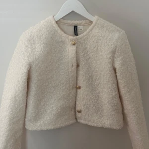 Beige teddyjacka / kofta - Säljer en supermjuk beige-vit teddyjacka / kofta från H&M Divided. Den har en rund halsringning och stängs med stiliga guldknappar framtill. Perfekt för att hålla sig varm och trendig!