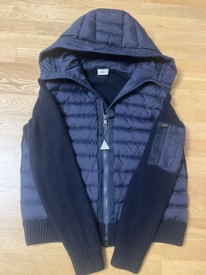 Mörkblå Moncler cardigan  - Snygg mörkblå pufferjacka från Moncler med stickade ärmar och huva. Jackan har dragkedja framtill, ribbade muddar och en ficka med dragkedja på ärmen. Klassisk Moncler-logga på axeln. Är i storlek L men passar även M, perfekt till alla vädrar och är i nytt skick.