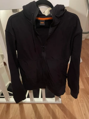 Svart hoodie från BOSS med dragkedja - Svart hoodie från BOSS med hel dragkedja framtill, huva med snörning och långa ärmar. Tröjan har en stilren look med diskret BOSS-logga på ärmen och praktiska fickor. Perfekt för chill dagar och enkel att matcha med jeans eller joggers.