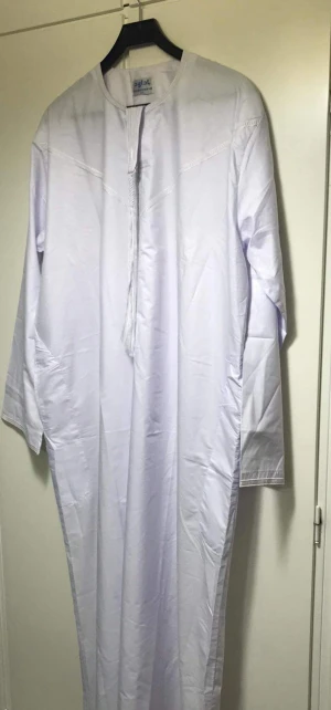 Vit långklänning från Badweed - Vit långklänning från Badweed med rund halsringning och dekorativ snörning framtill. Klänningen har lång ärm och är tillverkad i ett lätt bomullsmaterial. Enkel och stilren design med diskreta sömmar över bröstet och en rak passform.