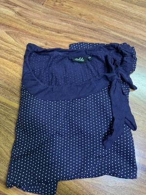 Mörkblå prickig blus med knyt från Isolde - Säljer en mörkblå blus från Isolde i storlek XXL med vita prickar över hela tyget. Blusen har rund halsringning och en snygg knyt-detalj vid axeln. Långa ärmar och mjukt, lätt material som känns skönt mot huden. Perfekt för dig som gillar klassiska mönster med en twist.