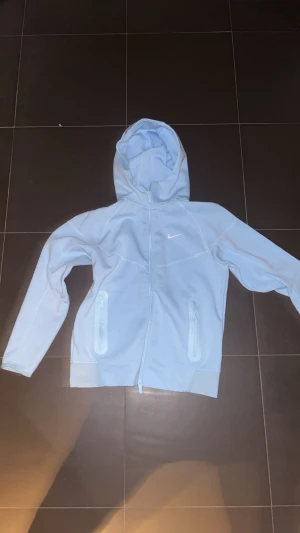 Nike NOCTA ljusblå hoodie M - Ljusblå hoodie från Nike NOCTA med hel dragkedja, huva och två fickor med dragkedja. Snyggt broderad Nike-logga på bröstet och coola detaljer på ryggen. Tillverkad i mjuk bomull och polyesterblandning, perfekt för chill eller träning. Det är dock ett litet fel på ena sidans dragkedja men inte märkbart Sålänge du inte har det uppzipad till toppen 