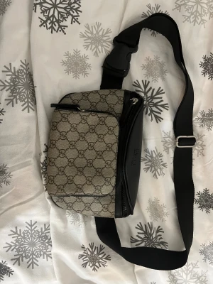 Gucci axelväska med monogram - Snygg axelväska från Gucci med klassiskt GG-monogram i beige och brunt. Väskan har svarta detaljer i skinn, justerbar axelrem och dragkedja framtill. Perfekt storlek för det viktigaste och riktigt trendig design.