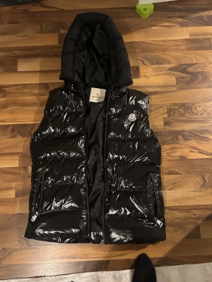 Svart dunväst från Moncler med huva - Snygg svart dunväst från Moncler med glansig finish och stor huva med Moncler-logga. Västen har två fickor med dragkedja och klassisk Moncler-märke på bröstet. Insidan har coolt serietryck och mönstrad etikett. Perfekt för dig som vill ha streetstyle med lyxkänsla.