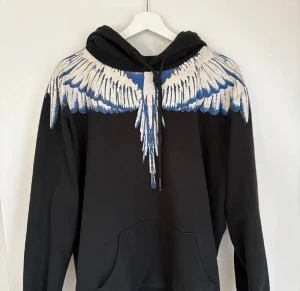 Svart hoodie med blått och vitt vingmönster - Säljer en svart hoodie från Marcelo Burlon med ett fett vingmönster i blått och vitt över axlarna och bröstet. Tröjan har huva med snörning och en stor magficka. Perfekt för dig som vill sticka ut med en unik design.