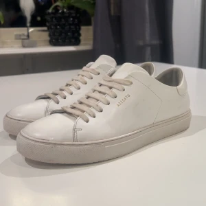 Vita sneakers från Axel Arigato - Snygga vita sneakers från Axel Arigato med minimalistisk design och diskret logga på sidan. Skorna har rund tå, platt sula och klassisk snörning. Tillverkade i skinn med en stilren look som passar till jeans eller dressade byxor.                                                   Pris kan diskuteras!