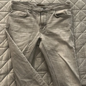 Grå jeans från Nudie Jeans, 33/32 - Snygga grå jeans från Nudie Jeans med klassisk femficksdesign och svarta broderade detaljer på bakfickorna. Jeansen har rak passform och är tillverkade i mjukt bomullstyg. Perfekta för dig som gillar stilrena och tidlösa jeans. Det är ett litet hål vid skrevet som ktnr syns på bild med kan få en bättre bild vid förfrågan! 