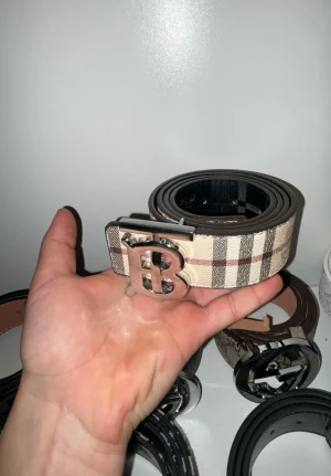 Rutig Burberry bälte med B-spänne - Säljer ett snyggt Burberry bälte med klassiskt rutigt mönster i beige, brun och röd. Bältet har ett stort, silvrigt B-spänne framtill som ger en lyxig vibe. Materialet är skinn och bältet är stilrent och passar perfekt till jeans eller kostymbyxor.