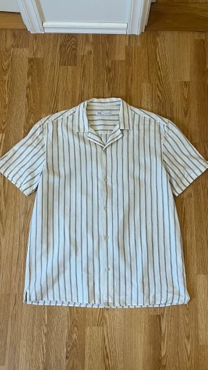 Randig kortärmad skjorta från Zara - Snygg kortärmad skjorta från Zara i storlek L. Skjortan är vit med tunna svarta vertikala ränder, har klassisk krage och knäppning framtill. Tillverkad i ett luftigt linnematerial som passar perfekt när det är varmt ute. Enkel och stilren design som är lätt att matcha.