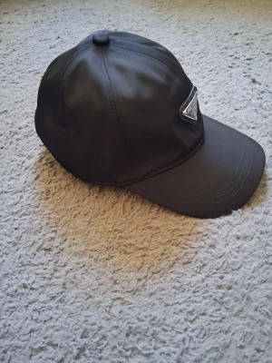 Svart Prada keps i nylon, storlek L - Snygg svart keps från Prada med ikonisk metalltriangel framtill. Tillverkad i återvunnen nylon (Re-Nylon) som ger en modern och clean look. Perfekt accessoar för dig som gillar exklusiva märken och streetstyle. Justerbar passform och coola detaljer på insidan.
