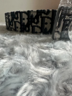 Svartvitt bälte  - Snyggt bälte med tydligt svartvitt mönster och ett stort, silverfärgat spänne framtill. Bältet har en bred design och är tillverkat i textilmaterial med -loggan över hela bandet. Perfekt för att lyfta vilken outfit som helst.