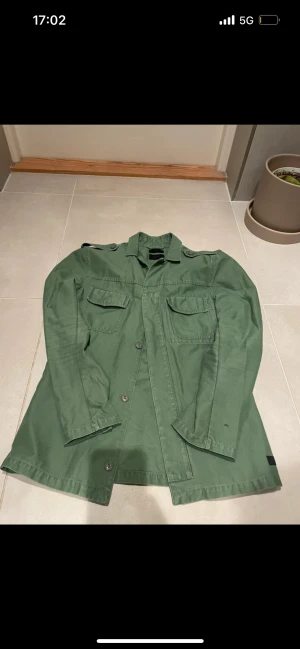 Grön overshirt med fickor - Snygg grön overshirt med klassisk krage och två stora bröstfickor med lock. Jackan har knäppning framtill med knappar och raka, långa ärmar. Materialet är bomull och modellen är normal i passningen. Perfekt att slänga över en hoodie eller t-shirt.