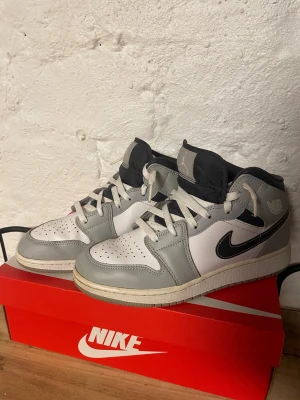 Nike Air Jordan 1 Mid grå/vit - Nike Air Jordan 1 Mid i en clean grå och vit färgkombination med svarta detaljer. Skorna har klassisk snörning, perforerad tå och ikonisk Jordan-logga på plösen. Ovandelen är i skinn och de har en platt sula. Perfekta för dig som gillar streetwear och sneakers.