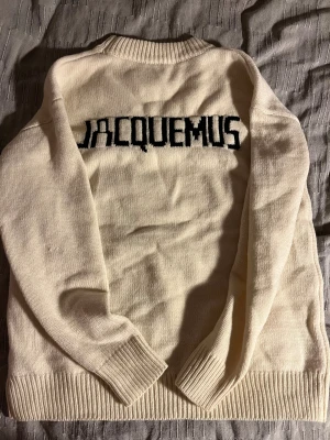 Vit stickad tröja Jacquemus - Säljer en clean vit stickad tröja från Jacquemus med svart logotyp på ryggen. Tröjan har ribbad krage, muddar och nederkant, samt långärmad passform. Perfekt för dig som gillar stilrena vibes och vill ha ett statement-plagg i garderoben. PRISET ÄR ABSOLUT INTE HUGGET I STEN!