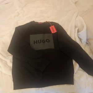 Svart sweatshirt från Hugo - Svart sweatshirt från Hugo med stor logga framtill i grått. Klassisk rund halsringning och långa ärmar. Perfekt för en clean och stilren look. Mjuk bomullskänsla och ribbade muddar vid ärmslut och nederkant. Helt ny med lapp kvar!