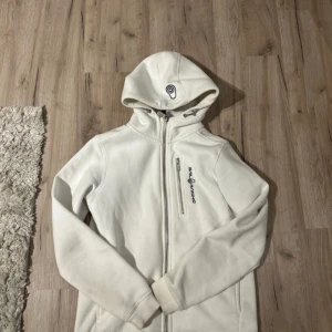 Vit sail racing kofta - Säljer en vit hoodie från sail racing med dragkedja framtill och en bröstficka med logga. Tröjan har huva med snörning och långa ärmar. Perfekt för kyliga dagar och enkel att matcha med det mesta.