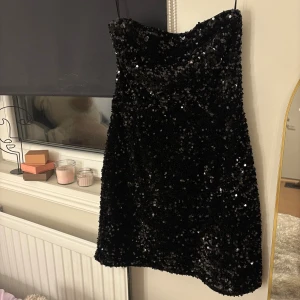 Svart paljettklänning axelbandslös nyår - Perfeeeeeekt nyårsklänning!!💘🥂 Enbart använd vid ett tillfälle. Axelbandslös svart kort klänning täckt av glittrande paljetter. Klänningen har en rak, något A-linjeformad siluett.