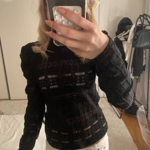 Svart spetsblus med lång ärm - Säljer en svart blus i spets med lång ärm och diskreta puffaxlar. Blusen har transparenta partier och fina detaljer i tyget, samt en liten knapp i nacken. Perfekt för dig som gillar en stilren men ändå edgy look.