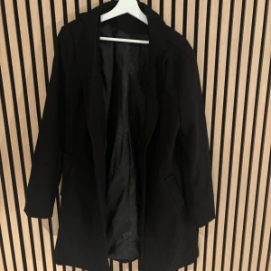 Svart klassisk kappa med knappar - Stilren svart kappa med klassisk krage och knäppning framtill. Jackan har två fickor på framsidan och även innerfickor, och dekorativa knappar vid ärmsluten. Perfekt för dig som gillar en clean och tidlös look. Fodrad insida för extra komfort. Storlek M
