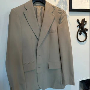 Beige kavaj från J.Lindeberg - 100% ull, Stilren beige kavaj från J.Lindeberg (storlek  46) med klassisk men lite LÄNGRE passform, VADDERADE AXLAR och snygga detaljer som spetsiga slag och flera fickor fram. (Fram fickorna är bara dekorativa). Fodret är glansigt och ger en lyxig känsla. Perfekt för dig som vill ha en clean och modern look.