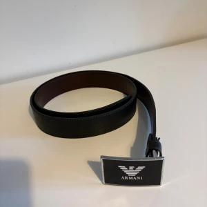Svart Armsn bäte - Snyggt svart bälte från Armani med en rektangulär spänne i silver och svart, prydd med den klassiska Armani-loggan. Bältet är tillverkat i skinn och har en stilren, slät design som passar till jeans eller kostymbyxor. Storleken på bältet är 130 cm och 4 cm bred.