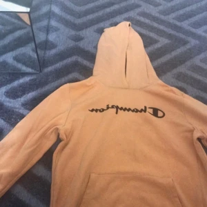 Beige Champion hoodie med svart tryck - Säljer en beige hoodie från Champion med svart logga över bröstet. Tröjan har huva och en stor magficka framtill. Perfekt för dig som gillar streetwear och vill ha en chill look.