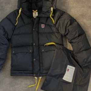 Svart Fjällräven Expedition dunjacka - Svart Fjällräven Expedition dunjacka med gula snören och huva. Jackan har tryckknappar med logga, stora fickor framtill och klassisk Fjällräven-märke på bröstet. Puffig och varm modell, perfekt för kalla dagar. Medföljer originalpåse med etikett. Storlek XS i herr storlek men passar mig som bär XS och S i damstorlek. Jag jämförde S i dam och XS i herr och de är snarlika. Nästintill helt oanvänd och super fint skick. Inga tecken på användning syns dvs inga fläckar, trasiga sömmar you name it. 