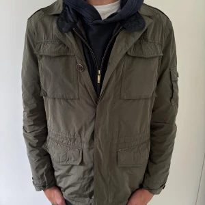 Olivgrön field jacket från Massimo dutti  - Snygg olivgrön parkajacka från Massimo dutti  med flera fickor och knappar på ärmarna. Jackan har en avtagbar quiltad innerjacka i mörkblått med dragkedja. Perfekt för lager-på-lager och har en clean, urban look. Passformen är normal och materialet känns lätt men slitstarkt.