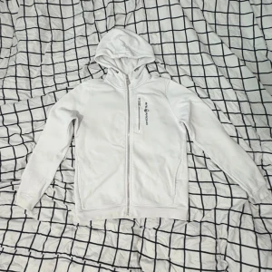 Vit hoodie från Sail Racing - Säljer en vit hoodie från Sail Racing med dragkedja framtill och en bröstficka med logga. Tröjan har huva, långa ärmar och är tillverkad i mjukt material som känns skönt mot huden. Perfekt för dig som gillar en clean och sportig stil.